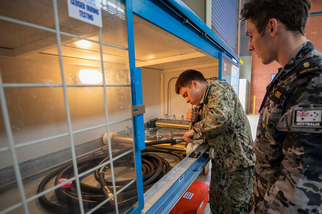 A sailor holds a hose aboard a ship as another watches.