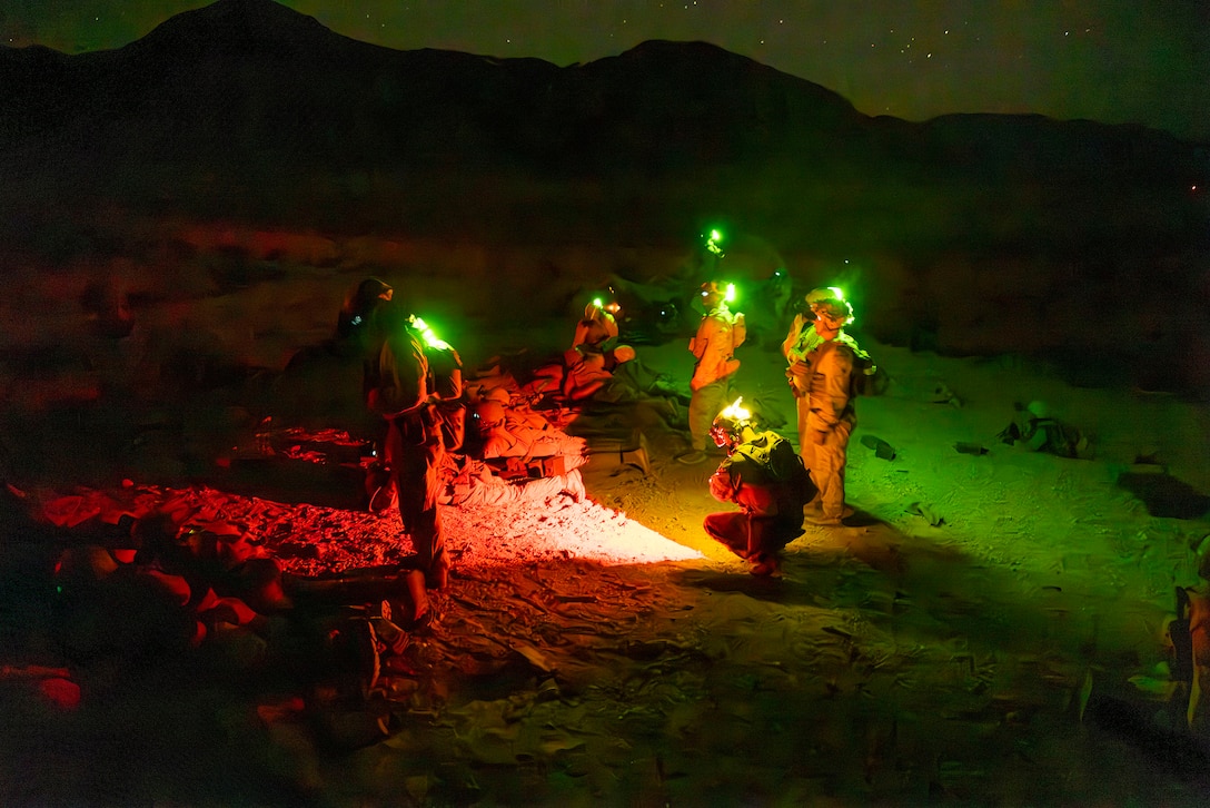 Marines wearing lights on their helmets stand in a desert area at night. They are illuminated by red and green lights.