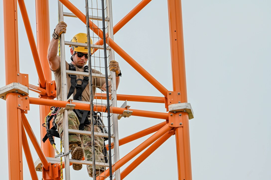 An airman wearing a helmet descends an orange communications tower during daylight.