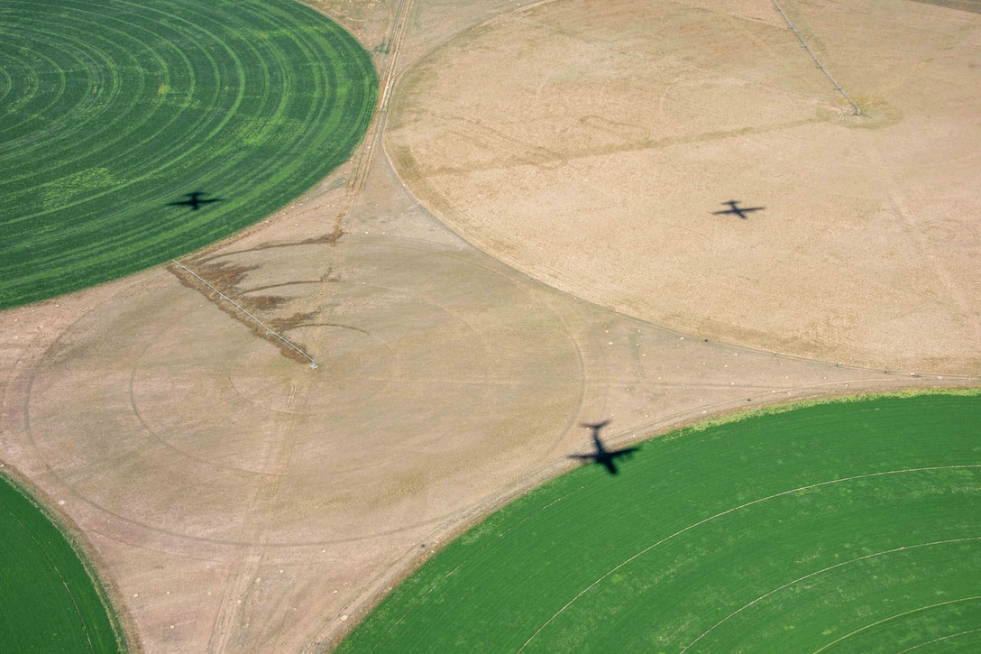 The shadows of three airborne aircraft are visible in a triangular formation on a concrete and turf surface.