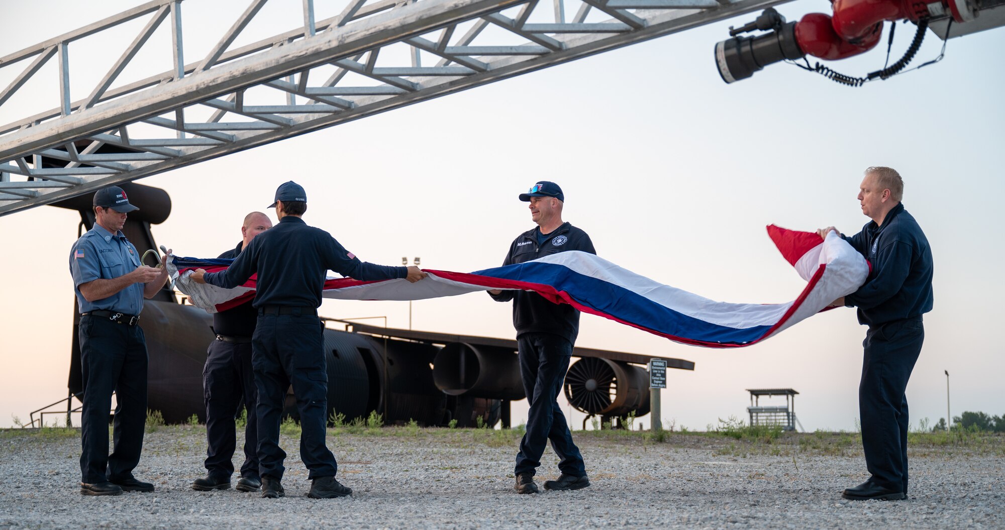 Five men unfolding a large American flag. In the foreground, at the top of the image, a ladder is seen partially. In the background, the metal frame of a fire training aircraft.
