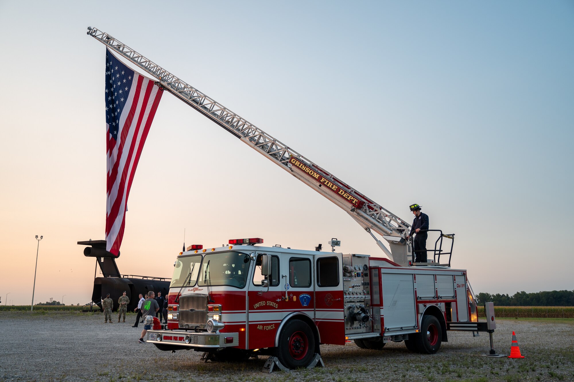 A fire truck hoisting a large American flag. On top of the truck, a firefighter. In the background, group of people standing around spectating and the metal frame of a fire training aircraft.