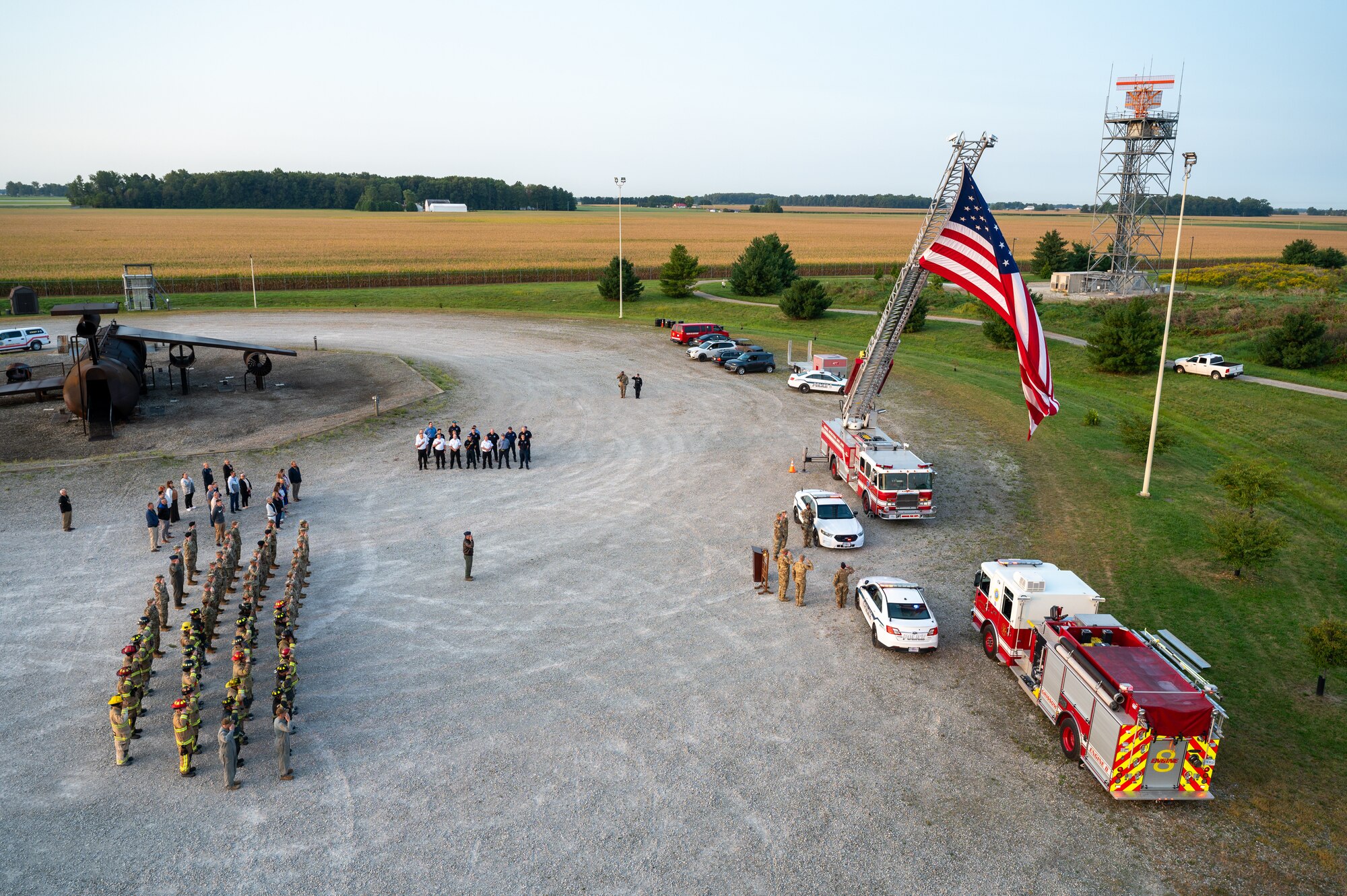 An aerial photo of an outside ceremony. A mid-sized military unit at left of the frame, various other groups scattered around them. At the right of the frame, various vehicles of fire responders, one fire truck hoisting a large American flag. In front of the vehicles, a podium and a few servicemembers are visible.