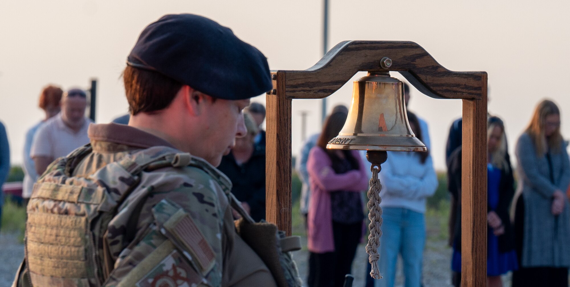 A woman in a U.S. Air Force uniform, with a black beret and vest on, bows her head. A bell located directly in front of her with the U.S. flag reflected inside of it.
