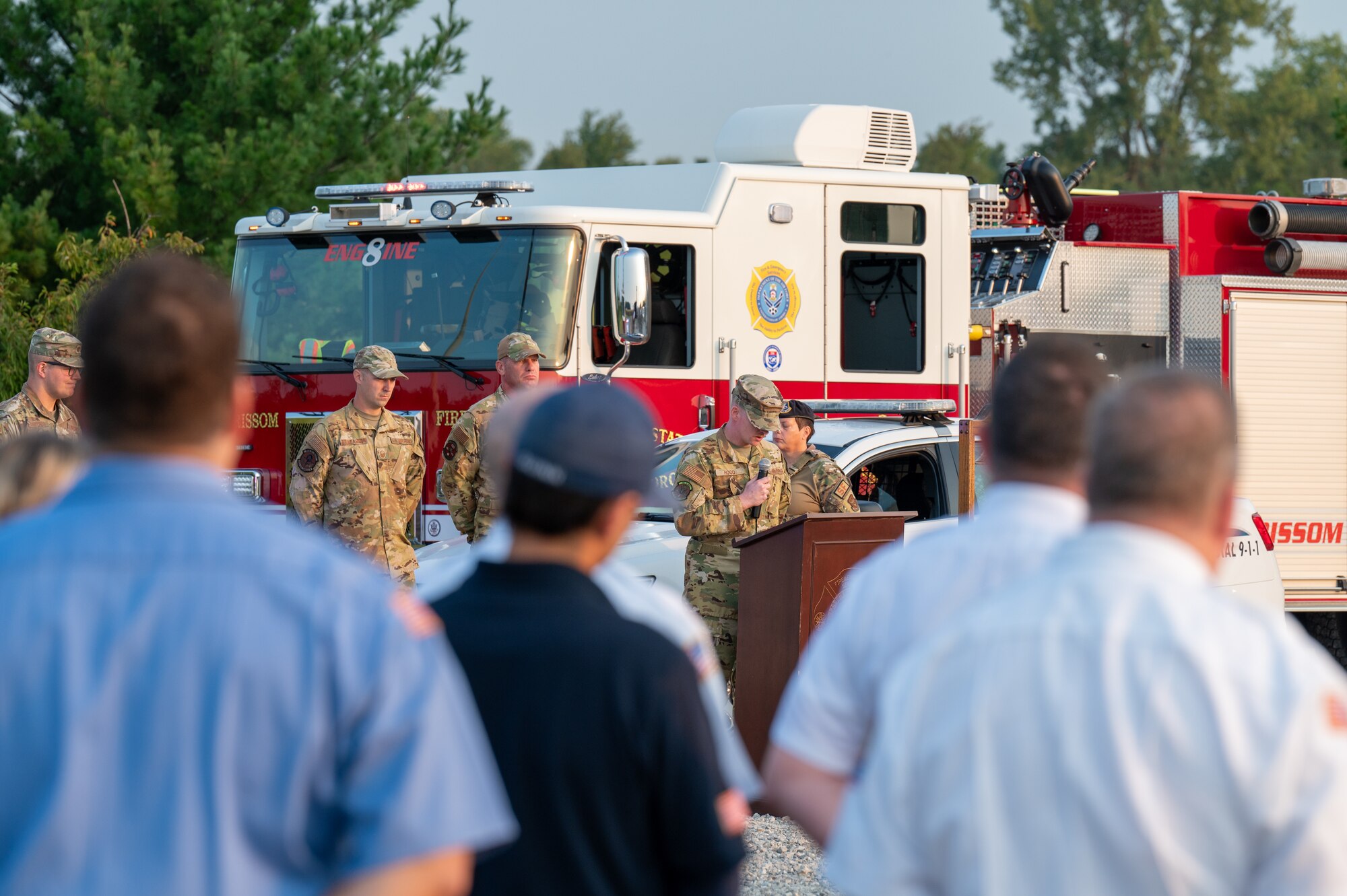 Men in short-sleeve firefighter uniforms look at a uniformed man speaking into a microphone behind a podium. Behind him, more servicemembers and a fire truck.