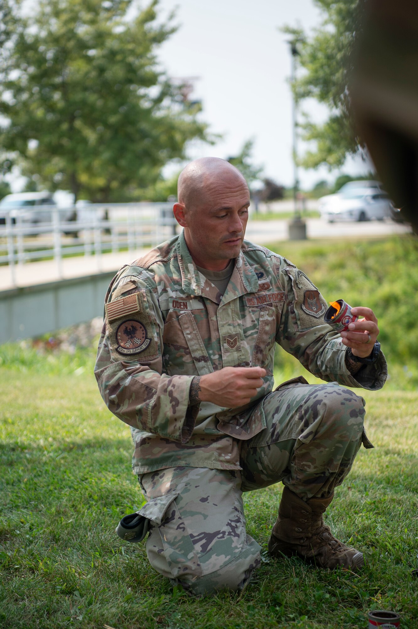 A man in a U.S. Air Force uniform kneels, holding a lit fire starter. In the background of the photo, a pedestrian bridge.