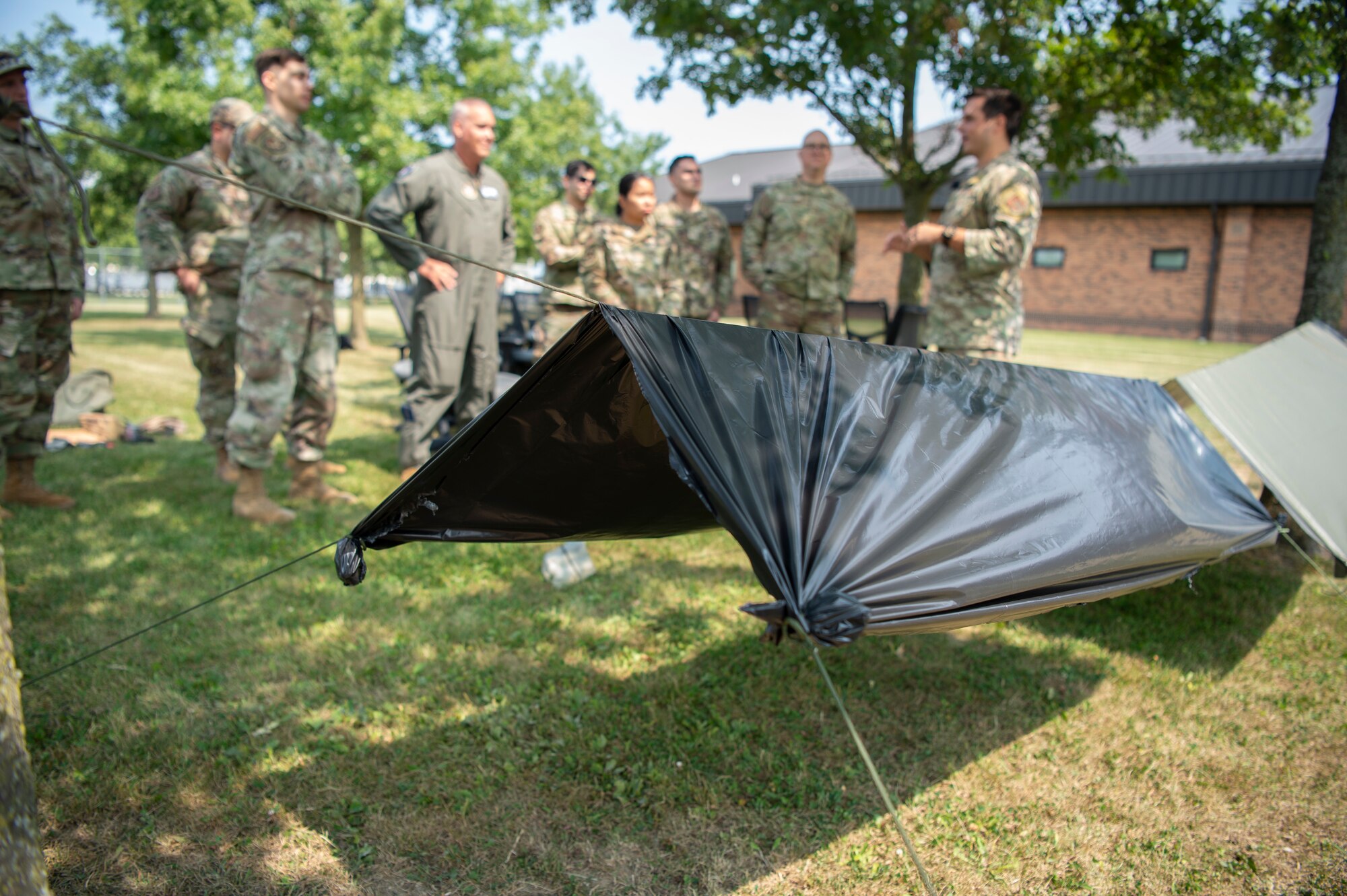 A man in a military uniform briefs other military members outside. In the foreground of the photo, tent-like shelters are seen hanging between trees.