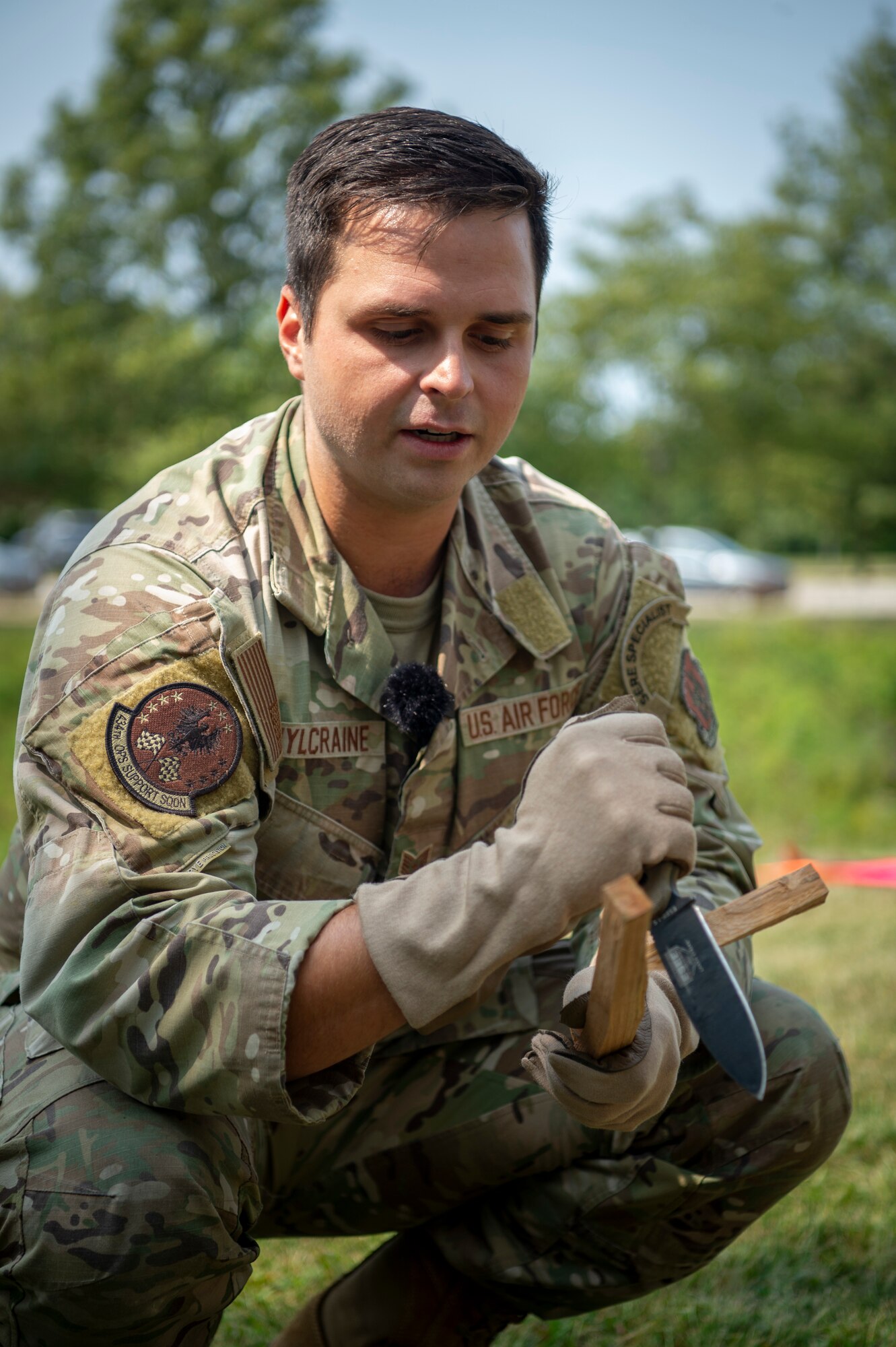A man in a U.S. Air Force uniform cuts a piece of wood while crouching outside. In his hand, a knife. In the other, a block of wood.