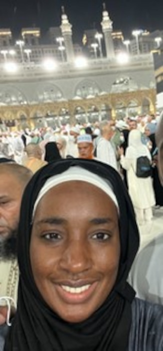 Woman in hijab, smiling at camera with a crowd of people behind her.