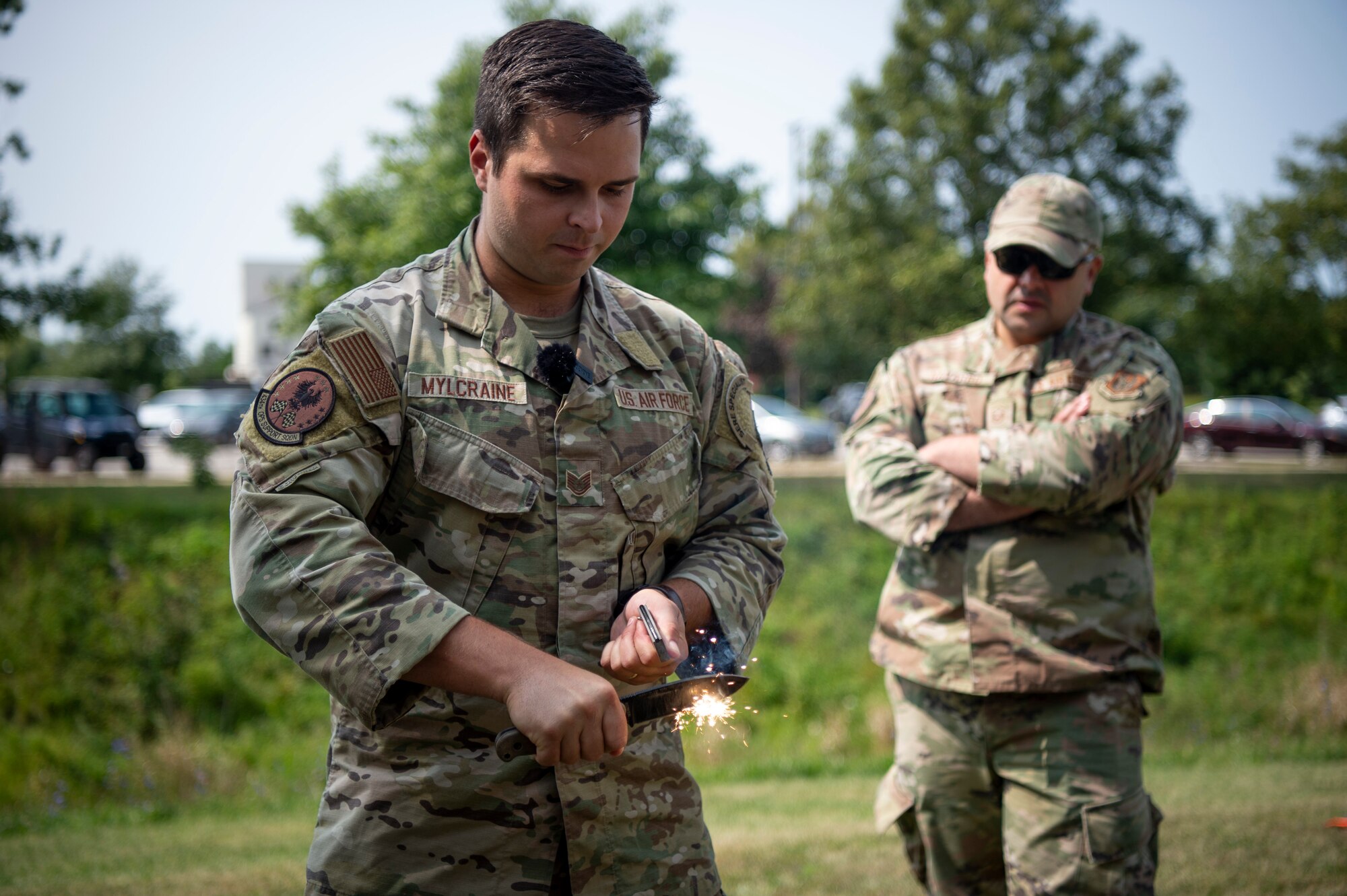 A man in a U.S. Air Force uniform creates sparks with a knife outside. Another uniformed man watches over his shoulder.