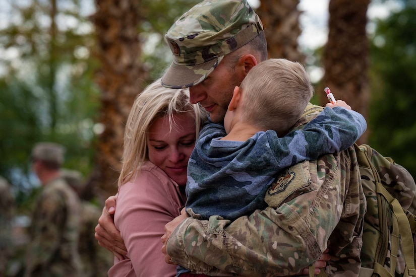 A man in a military uniform holds a small child in his arms, as a woman hugs him.