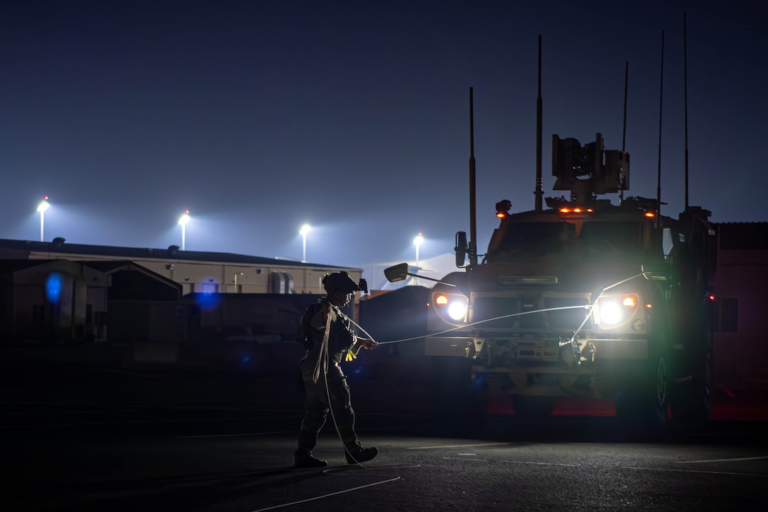 An airman carries a roll of cable that is partially on the ground and attached to a military vehicle while walking in a lighted area at night.