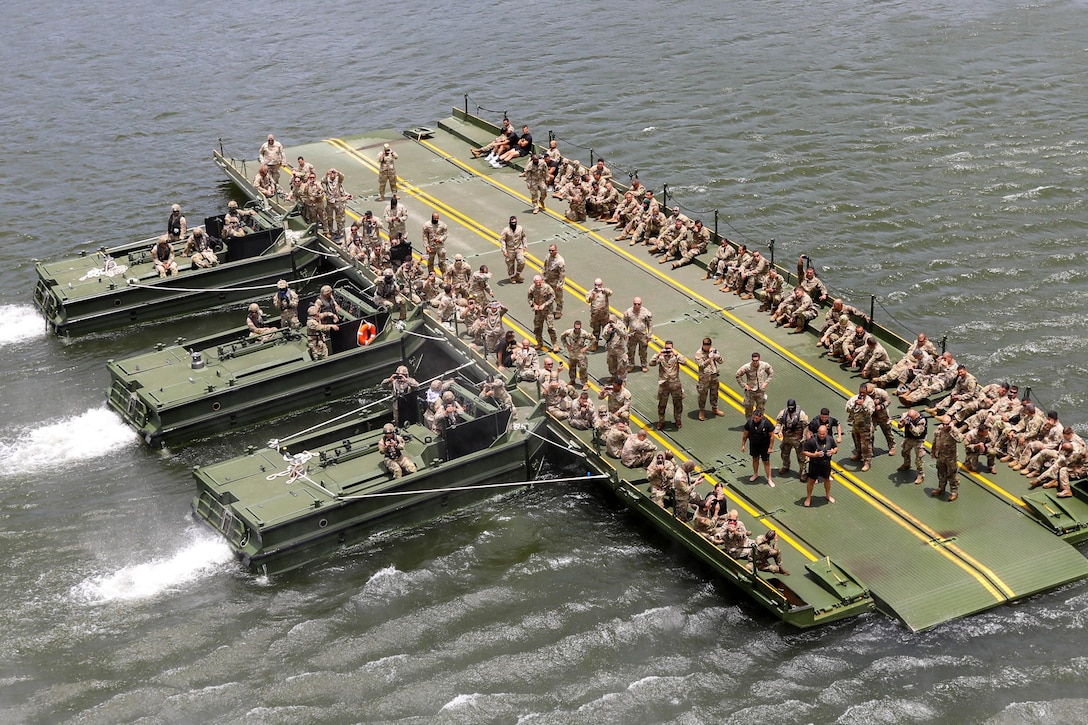 A large group of soldiers sit and stand on a mobile bridge being pushed through the water by three boats.