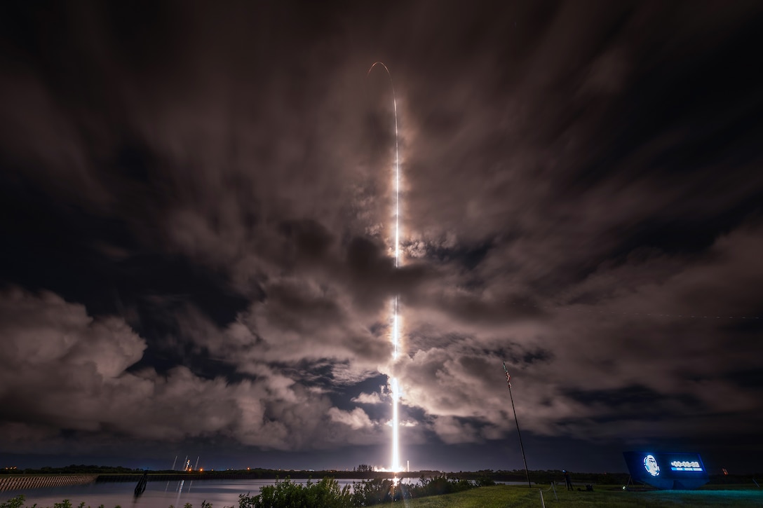 A rocket launches on a cloudy night, creating a bright white contrail in the sky.