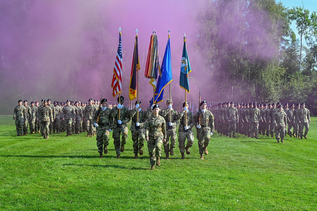 A color guard carries flags as rows of soldiers march in formation behind them on a grassy field during daylight as purple smoke fills the air.