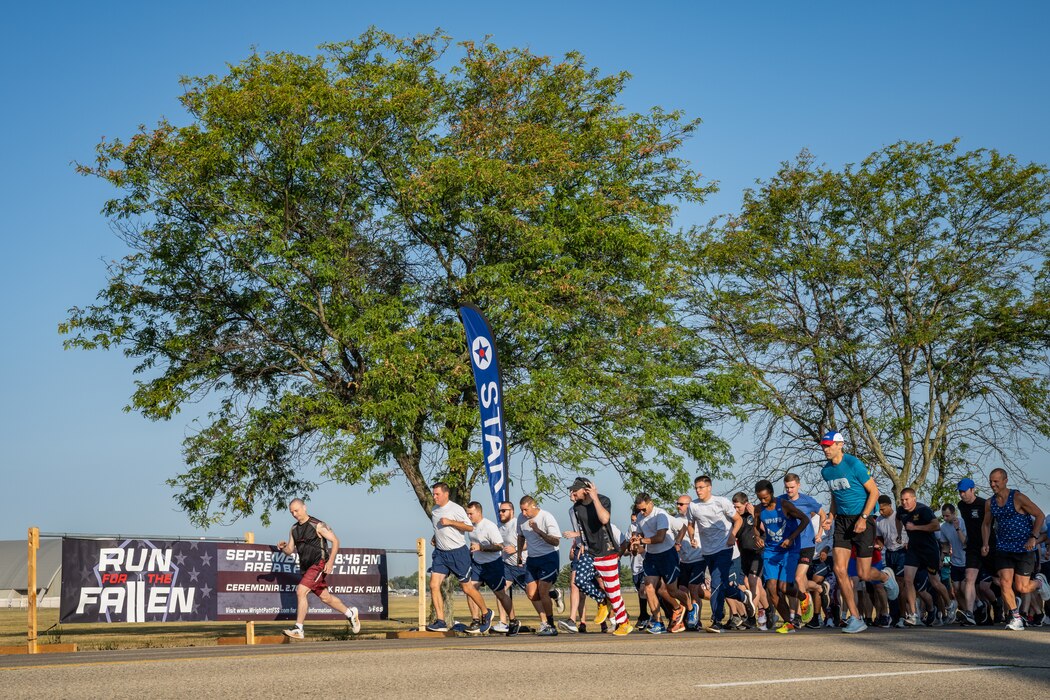 A large group of runners take off from a starting line beside a banner that reads Run for the Fallen and large trees