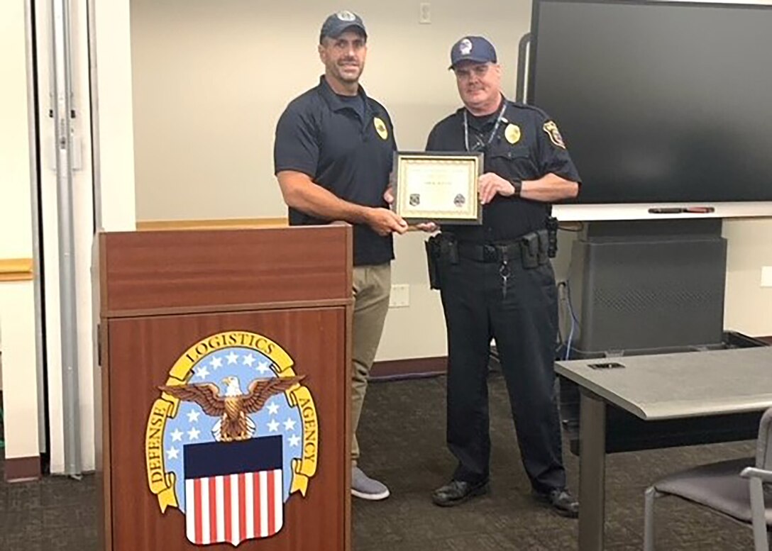 Two men stand shaking hands and holding a framed certificate.