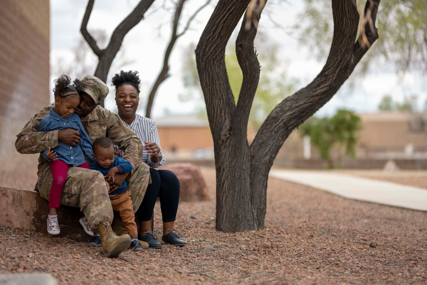A man in a military uniform sits on a step holding a toddler between his legs and a little girl on one leg. A woman watches from behind and laughs.