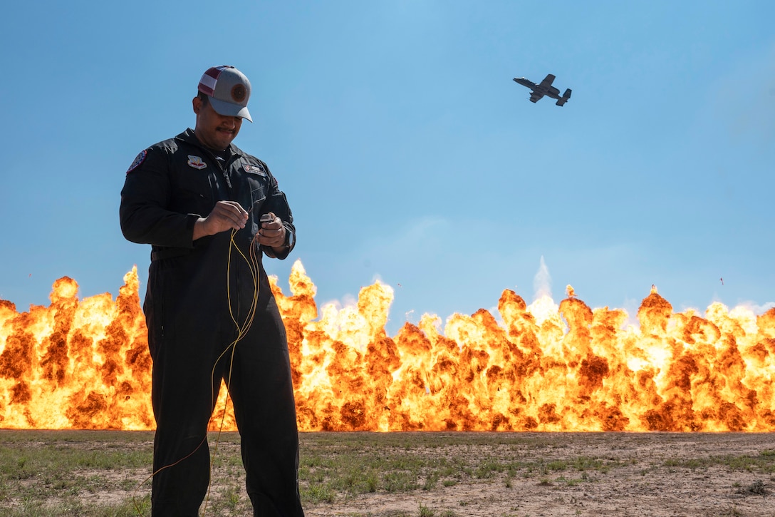 An airman connects wires to a device as large fire erupts in the background while an aircraft flies above.