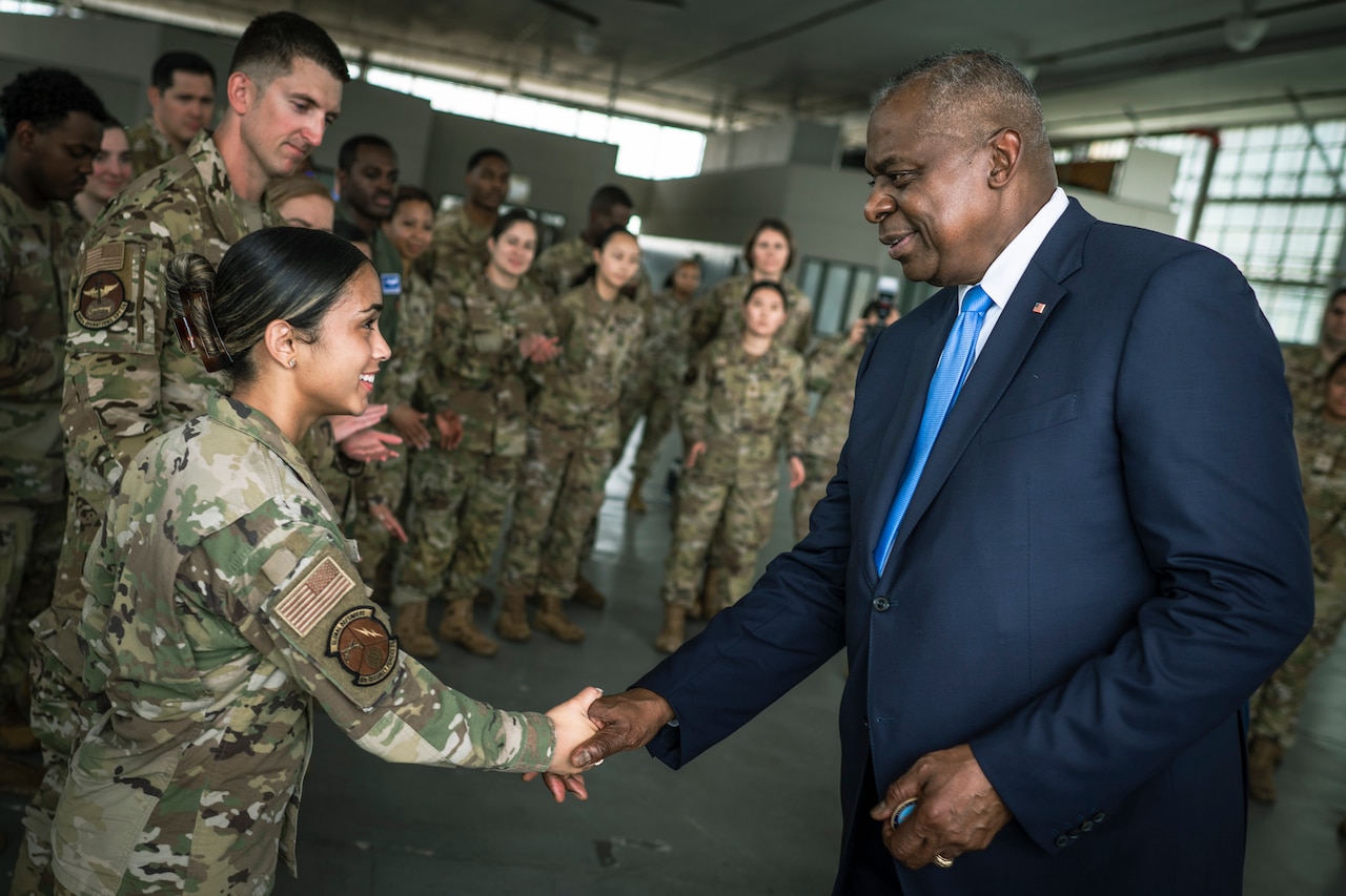 Secretary of Defense Lloyd J. Austin III shakes hands with a service member in a large room as other troops look on.