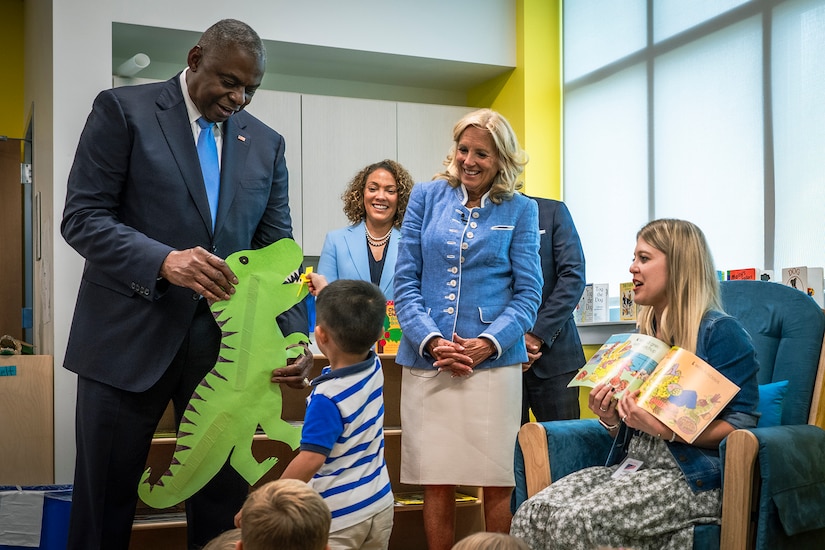 The defense secretary holds up a construction paper dinosaur and smiles at a child touching it as the first lady and others look on and smile.