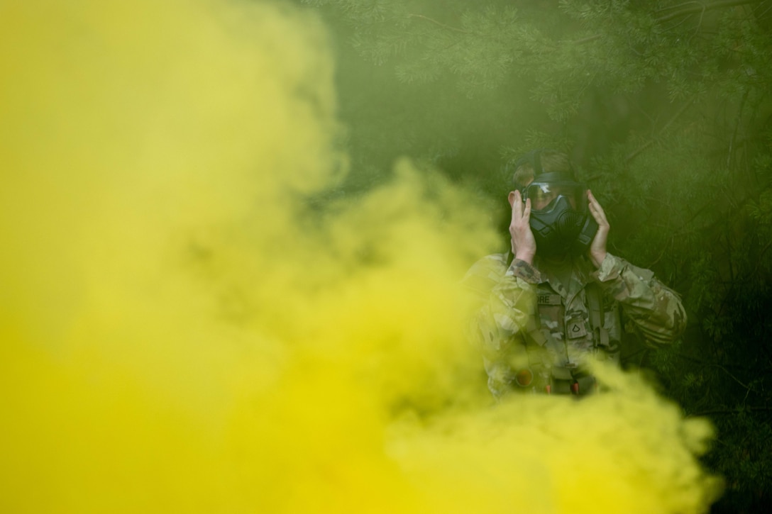 A soldier wearing a gas mask stands in a wooded area partially concealed by clouds of yellow smoke.