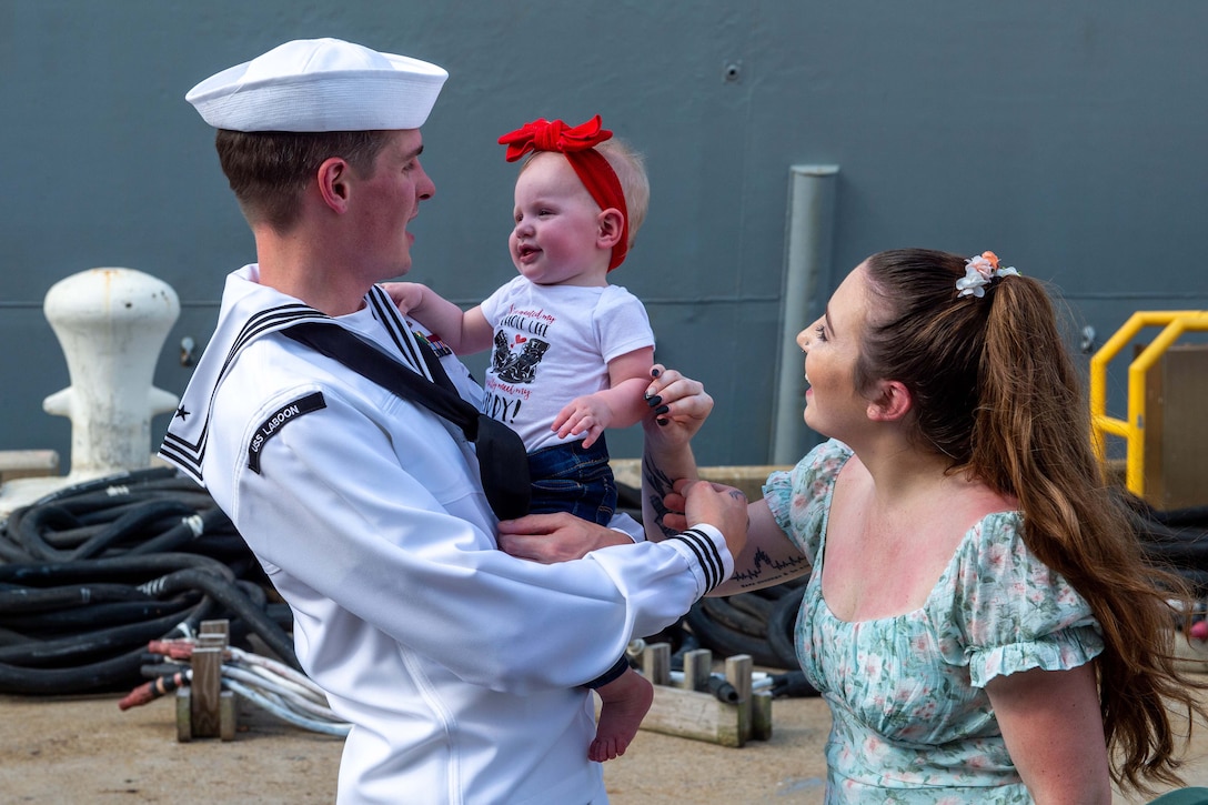 A sailor holds a baby as they look at each other while a civilian stands close by and looks at the same child.