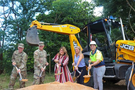 group of 5 people with shovels full of dirt posing for a ground breaking ceremony in front of a yellow backhoe.