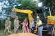 group of 5 people with shovels full of dirt posing for a ground breaking ceremony in front of a yellow backhoe.