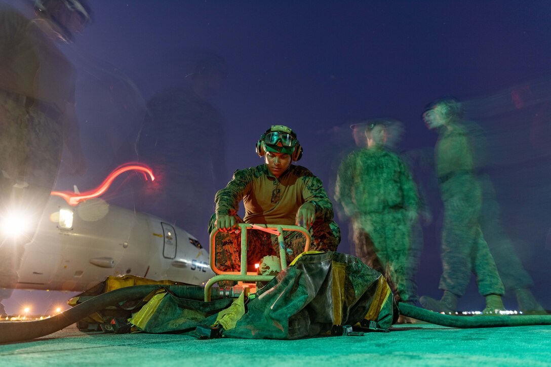 A Marine fuels a Navy aircraft at night with other Marines and a red light visible in the background.