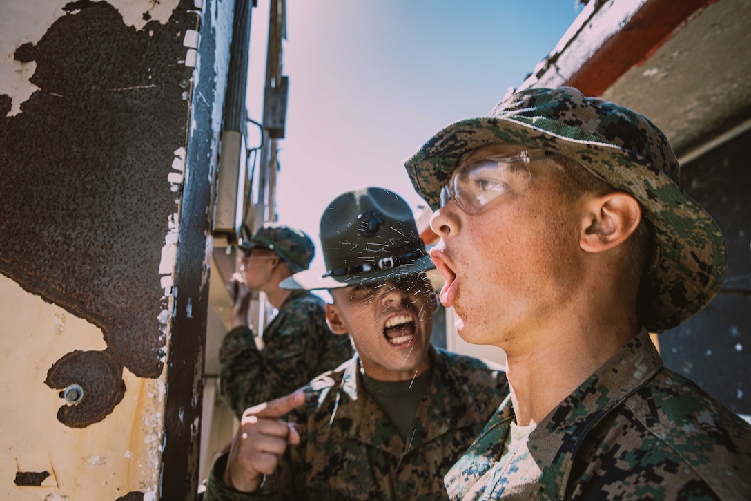 Spit flies as a drill sergeant points and shouts at a recruit in close proximity while another Marine is seen in the background.