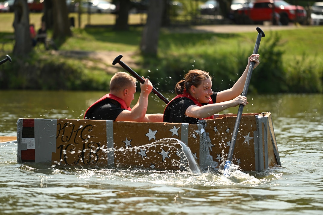 Water splashes as two soldiers use oars to row a makeshift boat in body of water with trees and grass in the background.