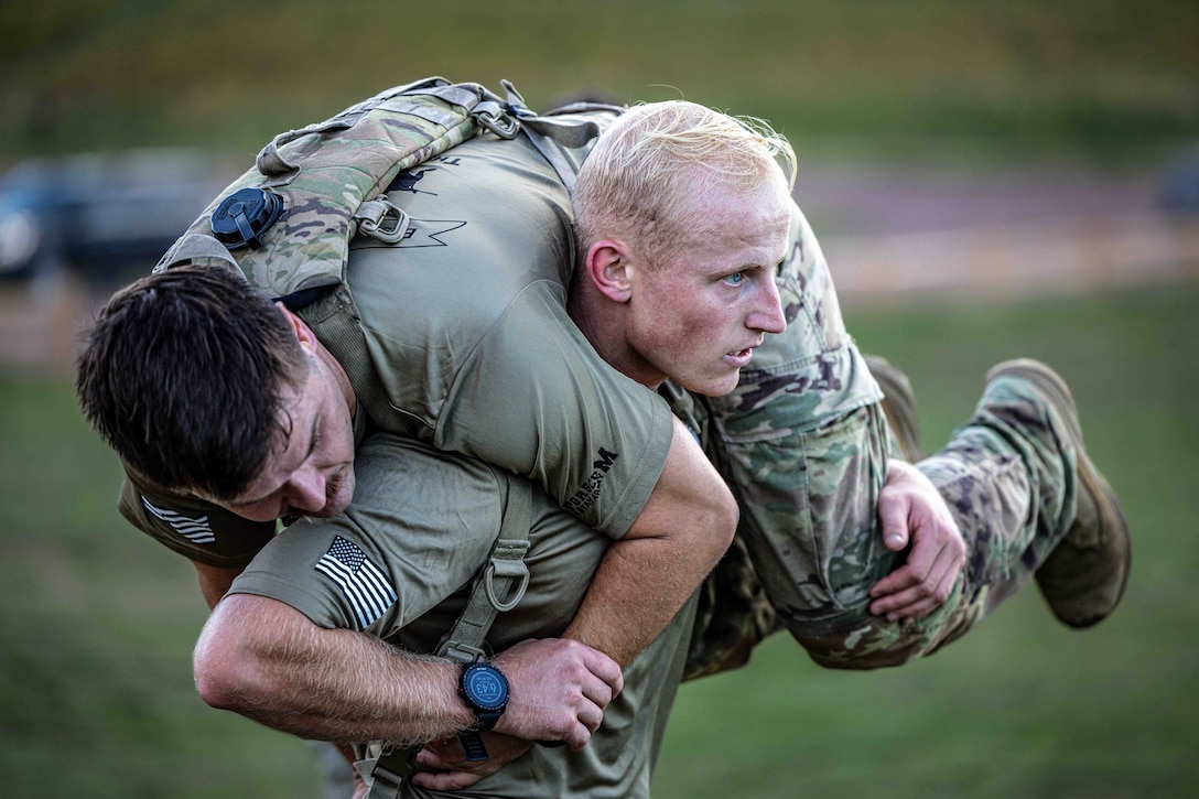 A soldier carries a fellow soldier on their shoulders while running in a field.