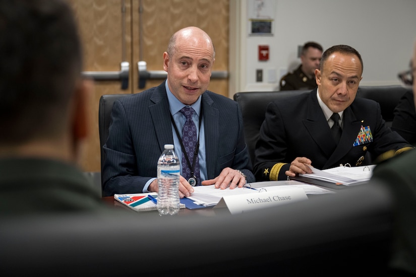 A person in a business suit is seated next to another person in a uniform.