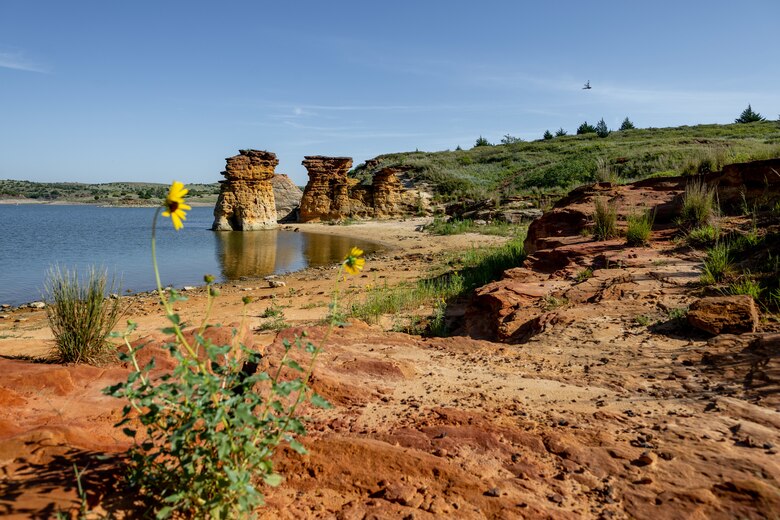 Large rock formations are in the background with water to the left, grass to the right, sky above, and red rock in the foreground with a yellow flower.