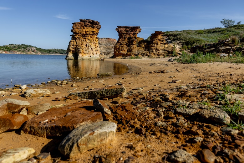 Large rock formations are in the background with water to the left, grass to the right, blue sky above, and red rock in the foreground.