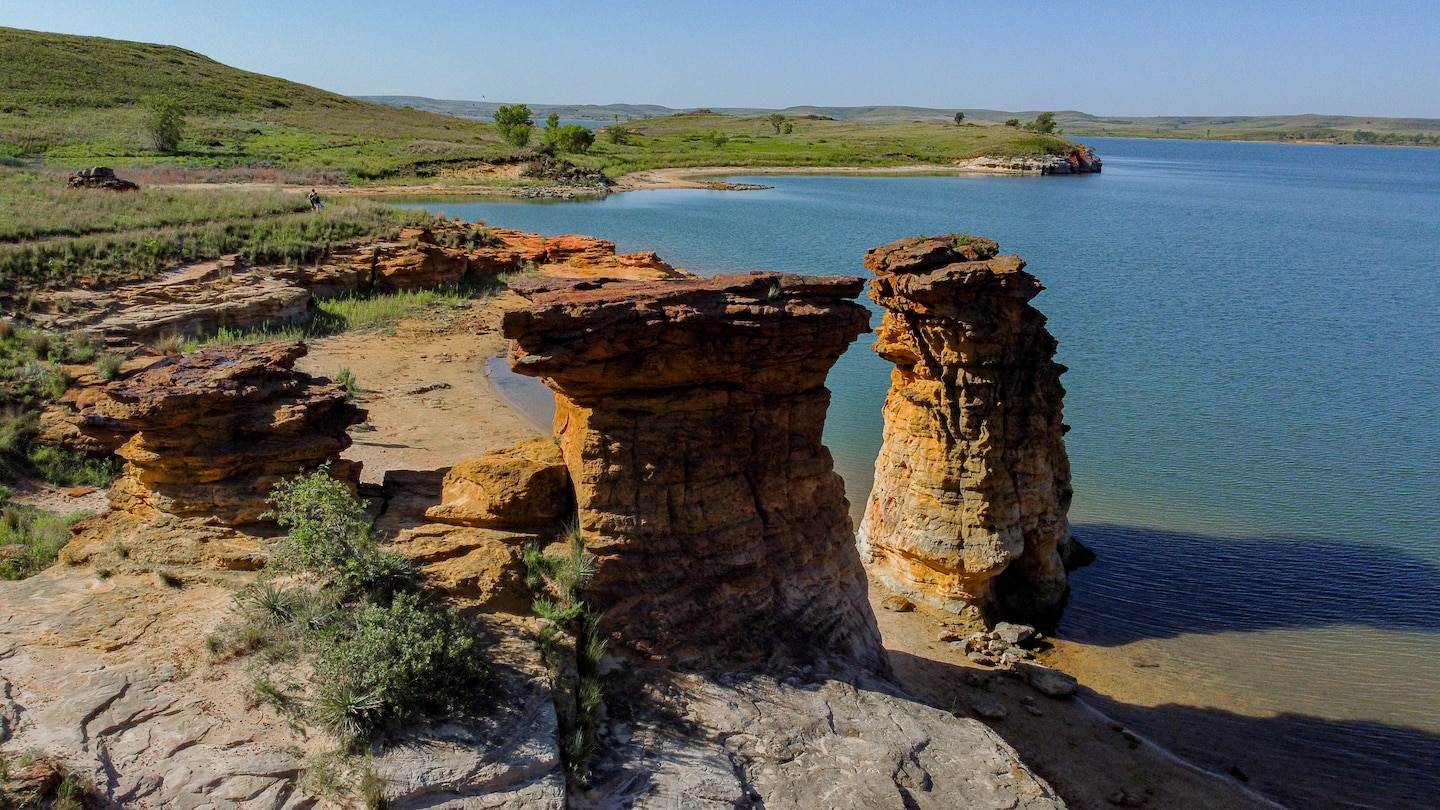 Two large rock formations stand with water to the right and grass to the left.