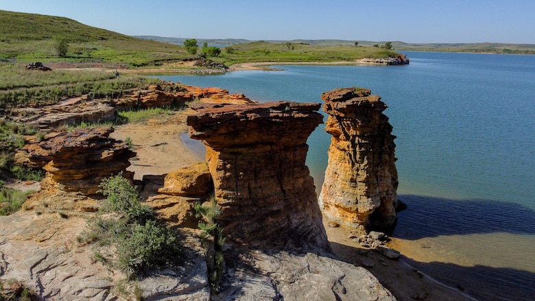 Two large rock formations stand with water to the right and grass to the left.