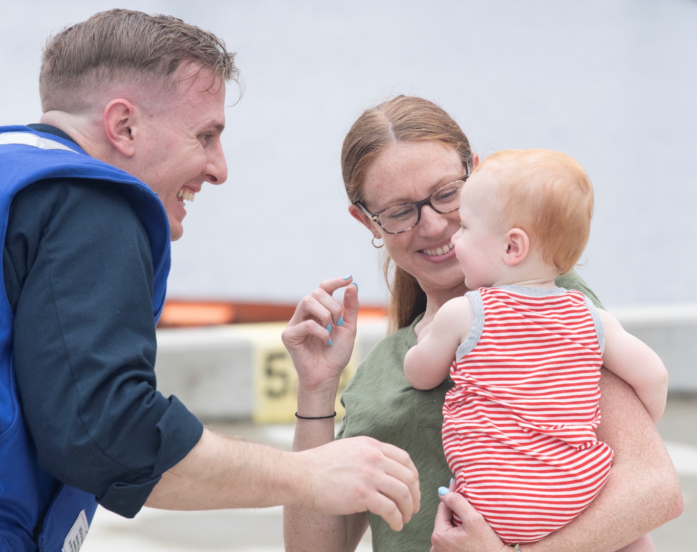 Smiling mother holds baby wearing onesie in her arm. Baby smiles up at grinning father.