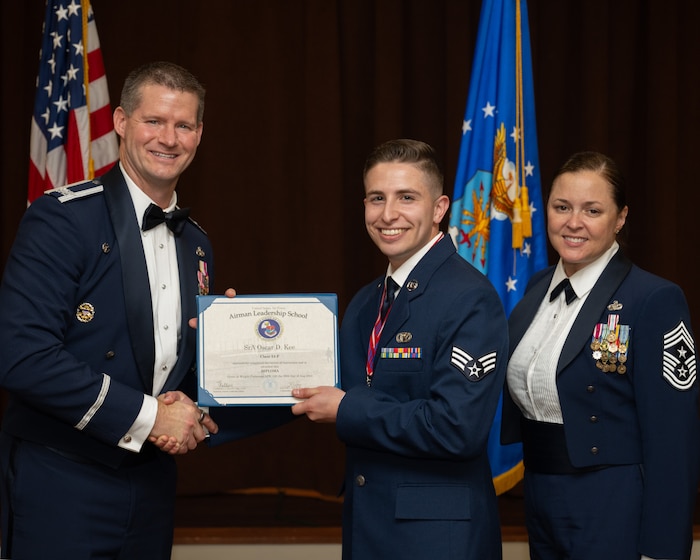 A male officer in Air Force Mess Dress uniform, on the left, hand a certificate to an enlisted man. A woman in a mess dress uniform stands on the right.
