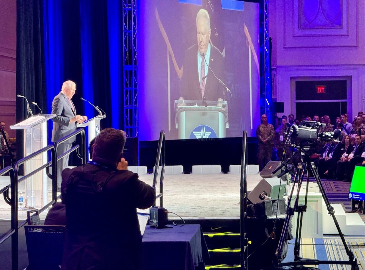 A man in a business suit stands at a lectern. There is a screen to the man’s left that is projecting a video of him at the lectern.