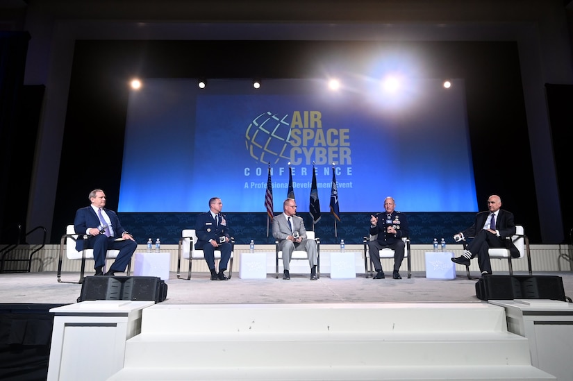 Five senior leaders sit on a stage during a panel in front of a large screen.