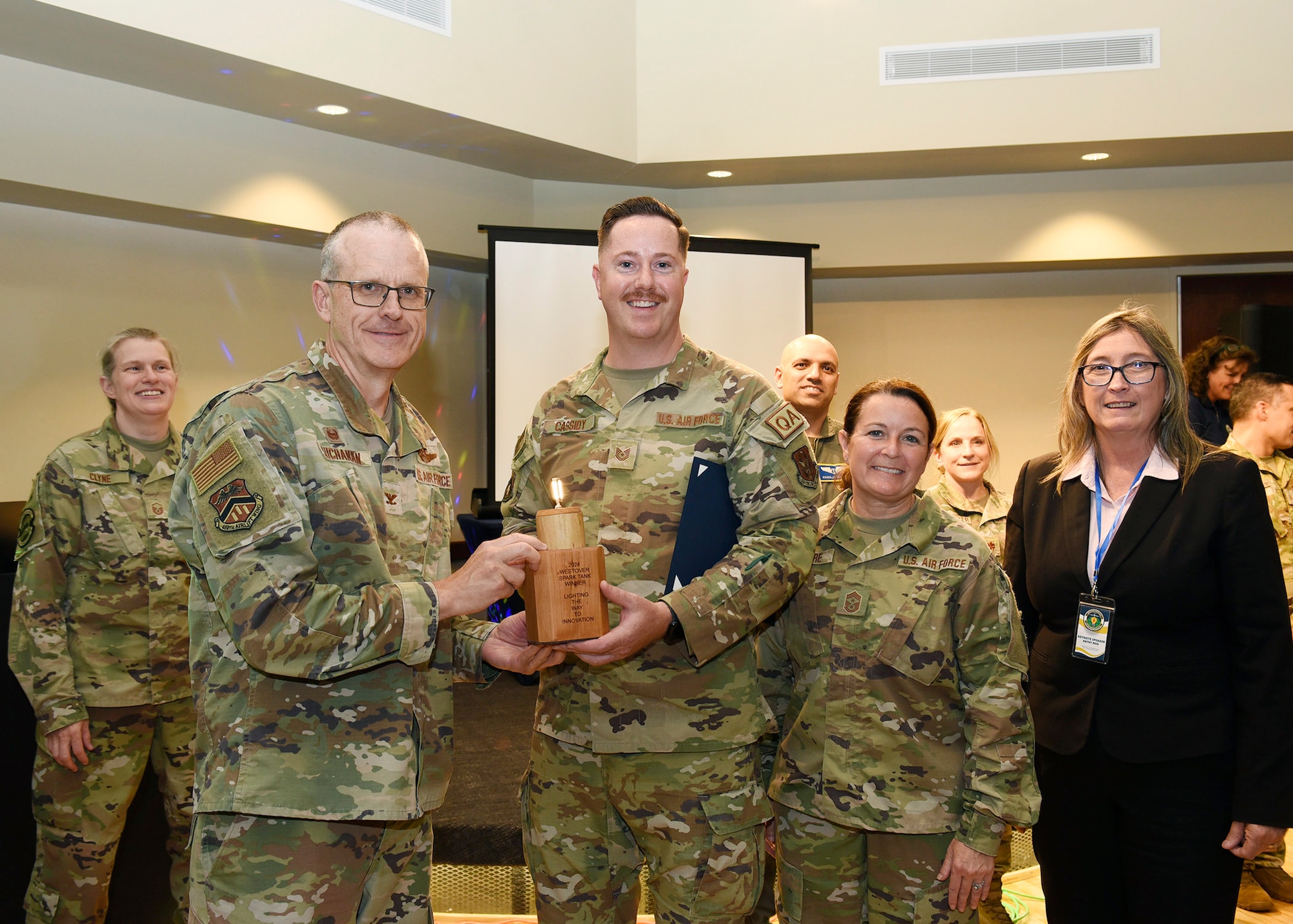 Tech. Sgt. Ryan Cassidy, 439th Maintenance Squadron’s Quality Assurance Section holds the winning trophy for the Spark Tank Challenge.  It is being handed to him by Colonel Greg Buchanan, 439th Airlift Wing commander.  Standing next to Cassidy is Chief Master Sgt. Barbara Gilmore, 439 AW command chief and Kathy Reid, AFWERX Spark division deputy.  The military members are in desert camouflage uniforms and Ms. Reid is in a black suit.  There are other military members standing in the background, and there is a white projection screen behind everyone.  Cassidy’s rescue hook was designed as a practical and efficient solution for rescuing individuals who fall from aircraft.