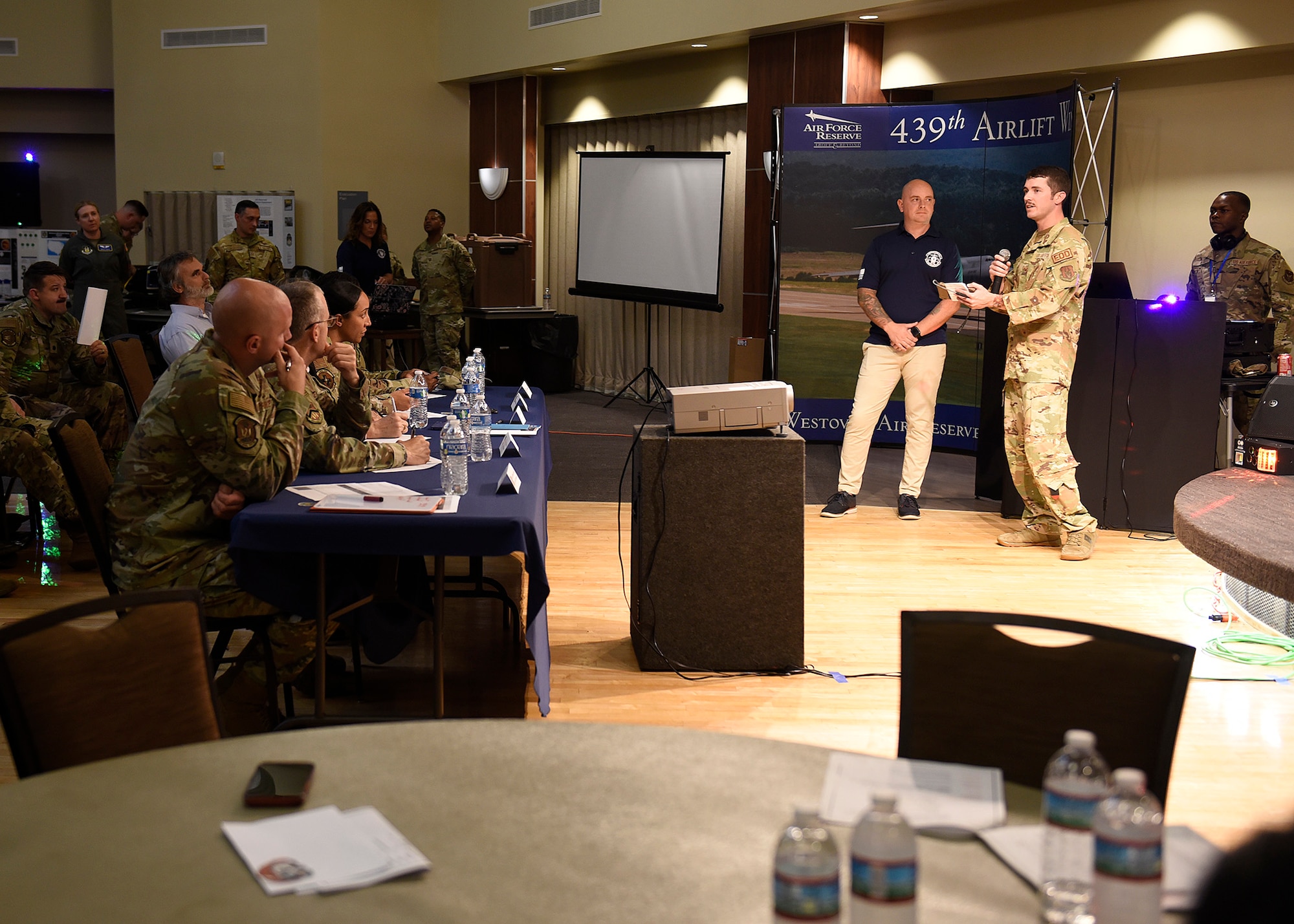 Staff Sgt. Brian Chamberlain, 439th Civilian Engineering Squadron’s Explosives Ordinance Section, pitches his innovation idea during the Spark Tank Challenge at Westover Air Reserve Base’s Club, Sep 6.  He is standing in front a projection screen, which shows information about his idea.  He is in a desert camouflage uniform, and standing next to a person in a blue polo shirt and tan pants.  They are facing the panel of judges during the presentation.