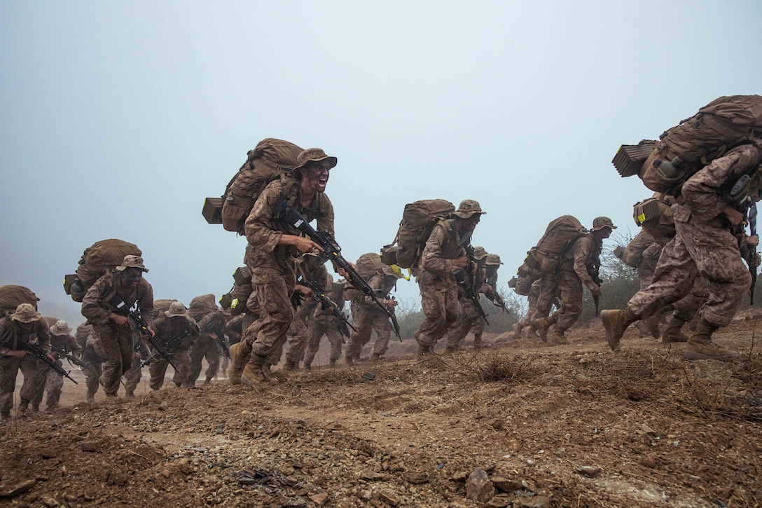 A large group of Marines carrying weapons and backpacks shout in unison as they climb up a dirt hill.