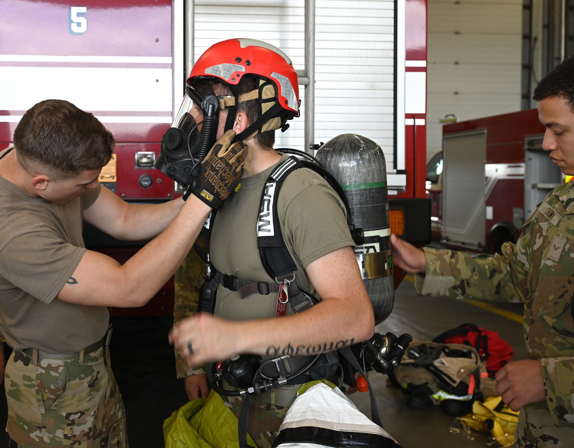 U.S. Air Force Airman Corban La Quey, center, 100th Civil Engineer Squadron Fire Department firefighter rookie, gets helped into his decontamination training suit as his breathing apparatus gets checked by fellow firefighters as part of the new “Rookie Academy” program at Royal Air Force Mildenhall, England, Aug. 27, 2024. The Rookie Academy is an intense training program put together by Staff Sgt. Connor Nance, 100th CES Fire Department assistant chief of training, and taught by multiple experienced firefighters. It condenses four months of training for new firefighters into three weeks. As part of the program, new rookie firefighters learn equipment use and life-saving techniques the same way, rather than being split with different trainers. (U.S. Air Force photo by Karen Abeyasekere)