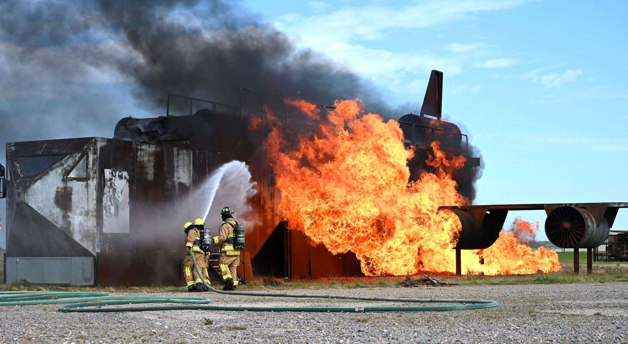 U.S. Air Force Airman Corban La Quey, left, and Airman Jeniel DeJesus, center, 100th Civil Engineer Squadron Fire Department firefighter rookies, spray water onto flames on a mock aircraft live fire trainer, backed up by Daniel Hodkinson, 100th CES Fire Department British civilian firefighter, at the end of their “Rookie Academy” course on Royal Air Force Mildenhall, England, Aug. 28, 2024. The Rookie Academy is an intense training program put together by the RAF Mildenhall Fire Department, condensing four months of training for new firefighters into three weeks. (U.S. Air Force photo by Karen Abeyasekere)