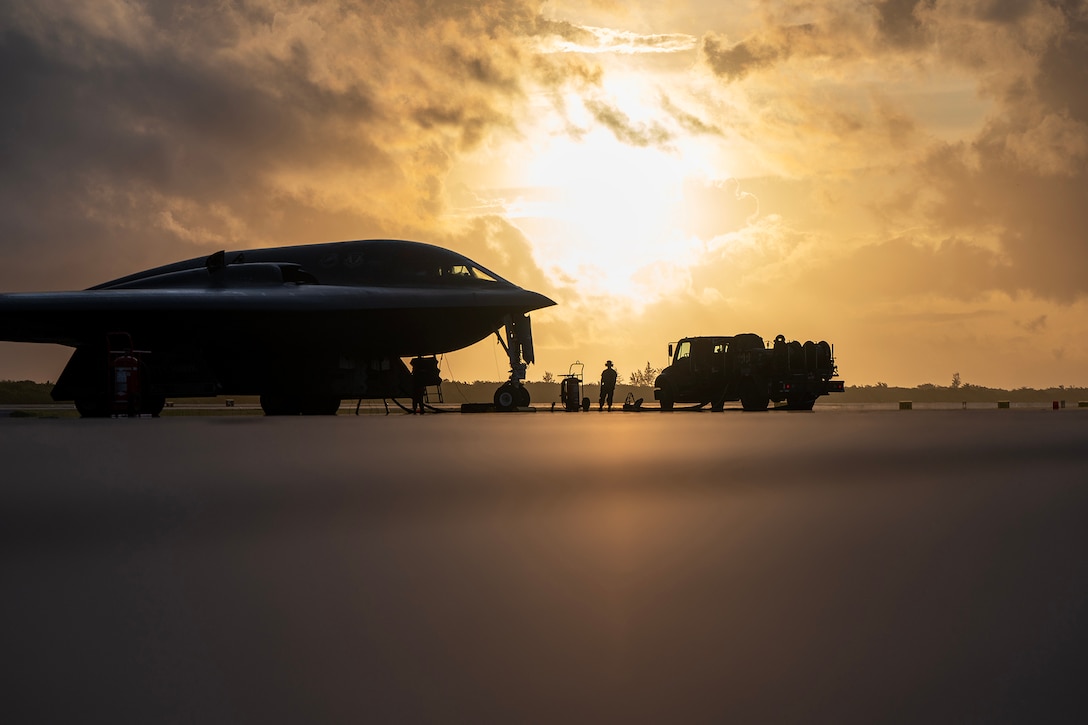 Airmen are shown in silhouette on a tarmac refueling a military aircraft at twilight with a low sun in the background.