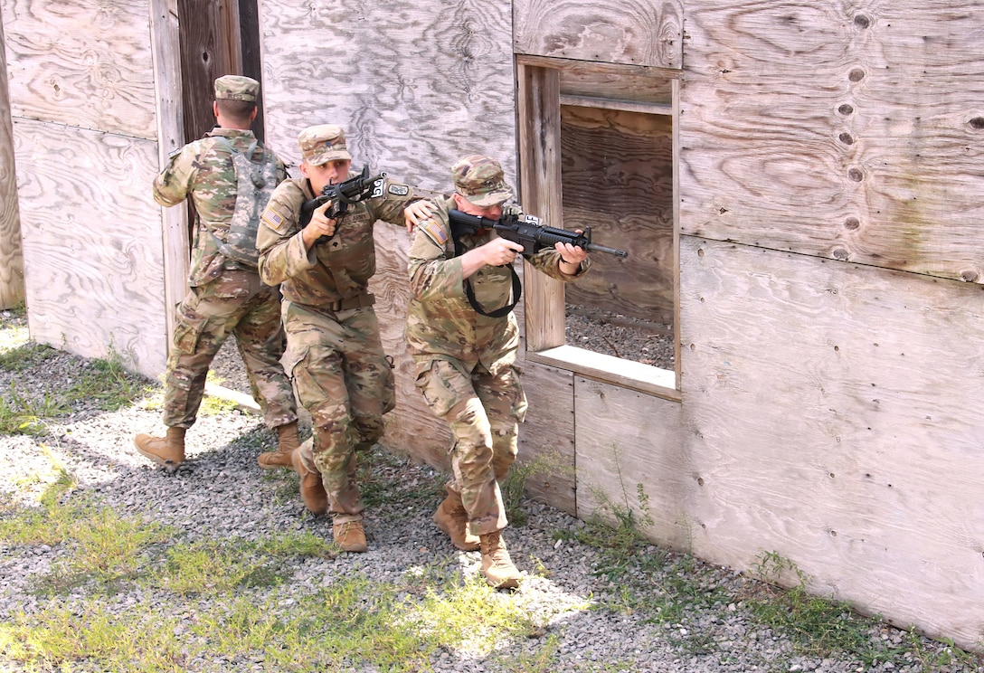 A soldier enters a wooden structure as two others walk in the opposite direction with their weapons drawn during a daylight competition.