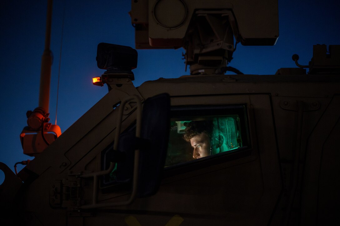 An airman sits inside a military vehicle while looking down at a screen at night with red, yellow and green lights illuminating the vehicle.